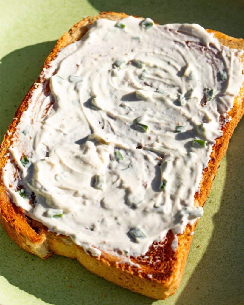 A slice of bread with cream cheese spread over in a sunny photo on a green plate.