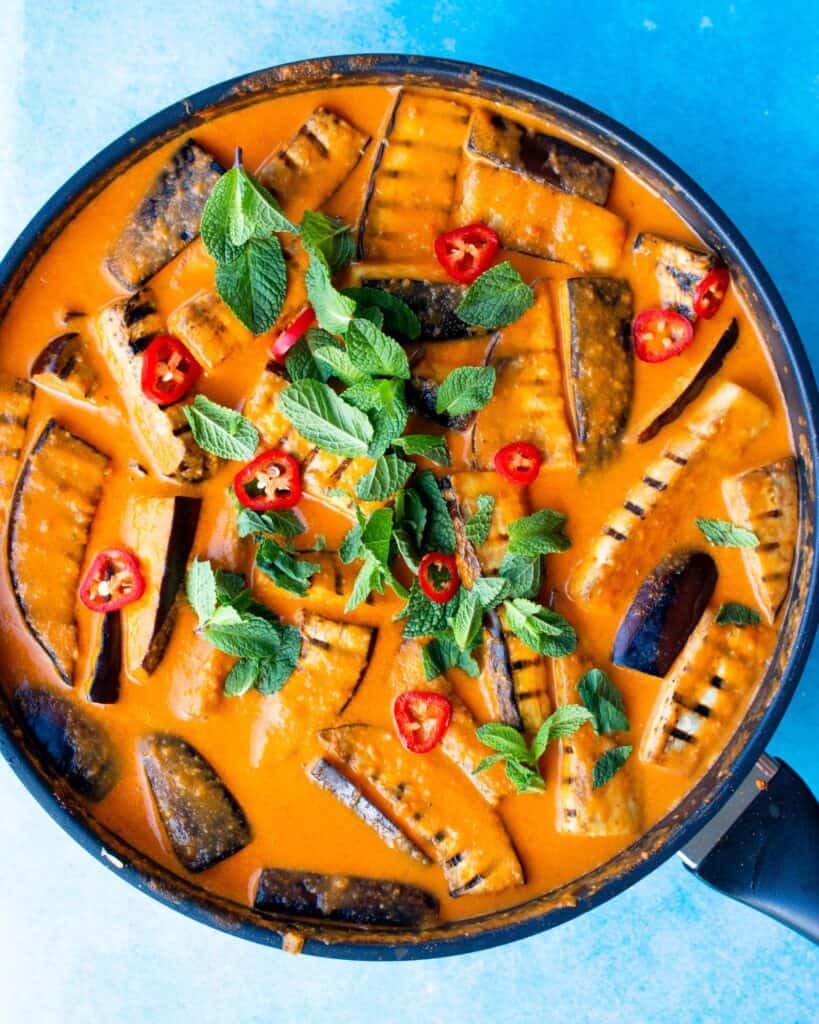 Aubergine curry in a large pan with aubergine charred batons covered in sauce topped with coriander and chillies.