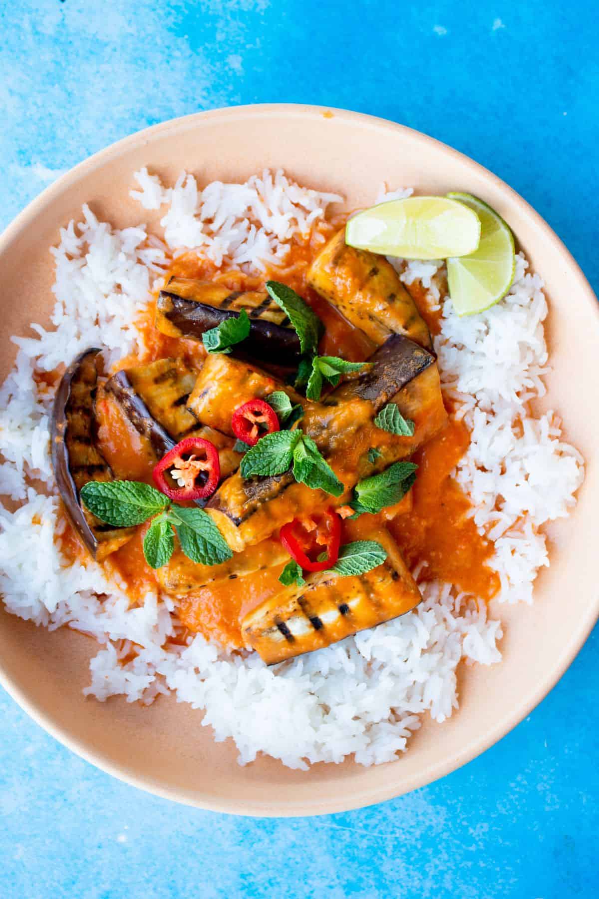 Aubergine curry served up in a bowl with white rice, mint, red chilli slices and lime wedges on a blue background. 