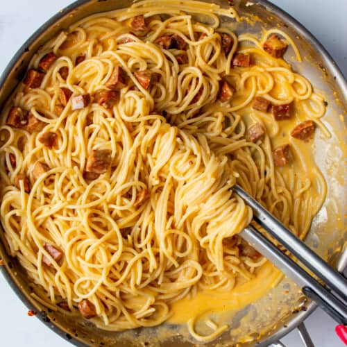 Chorizo carbonara with spaghetti, small slices of chorizo and carbonara sauce in a large stainless steel pan mixed with large metal tongs.