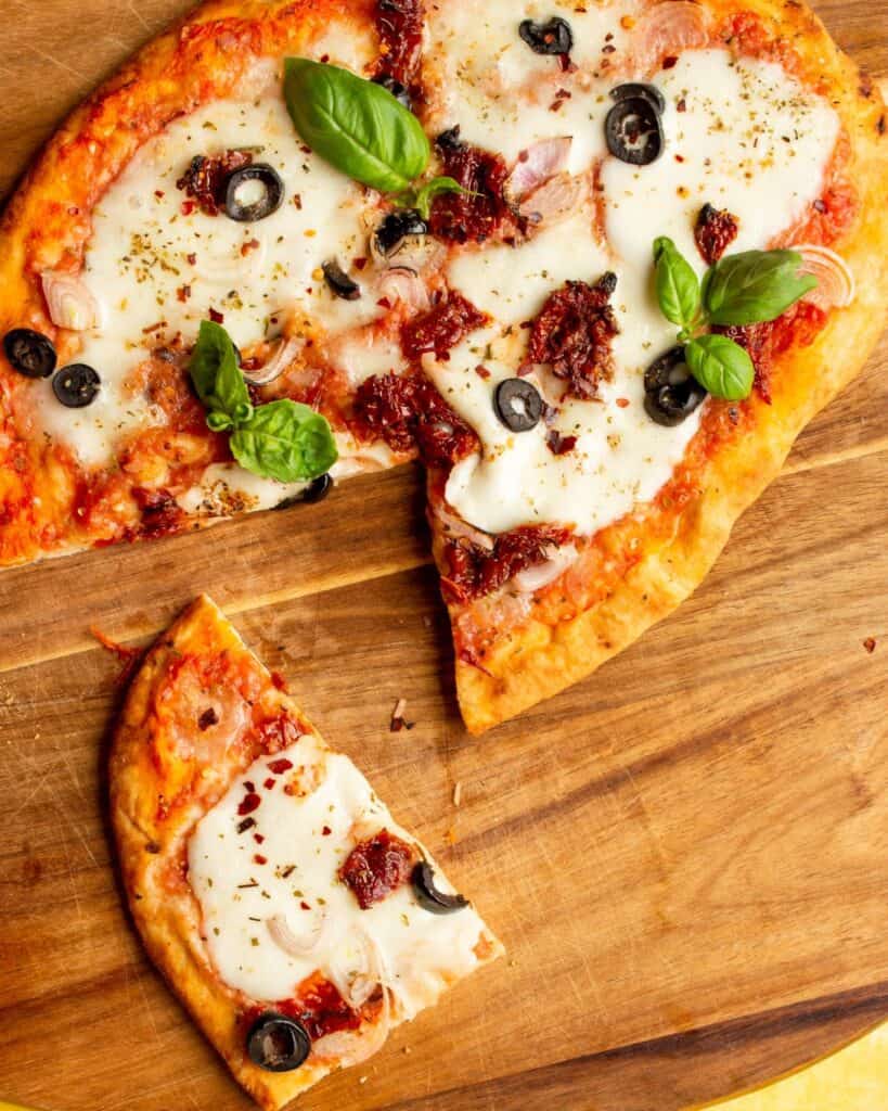 Naan bread pizza with mozzarella, sundried tomatoes and basil on a wooden board with one slice cut out on wooden board.