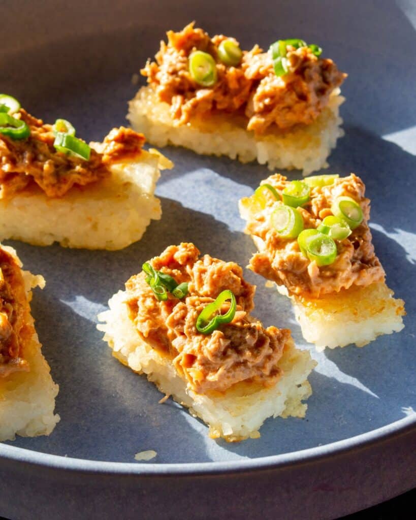 Rectangular pieces of rice topped with tuna and spring onion on a blue bowl in the sun shine.
