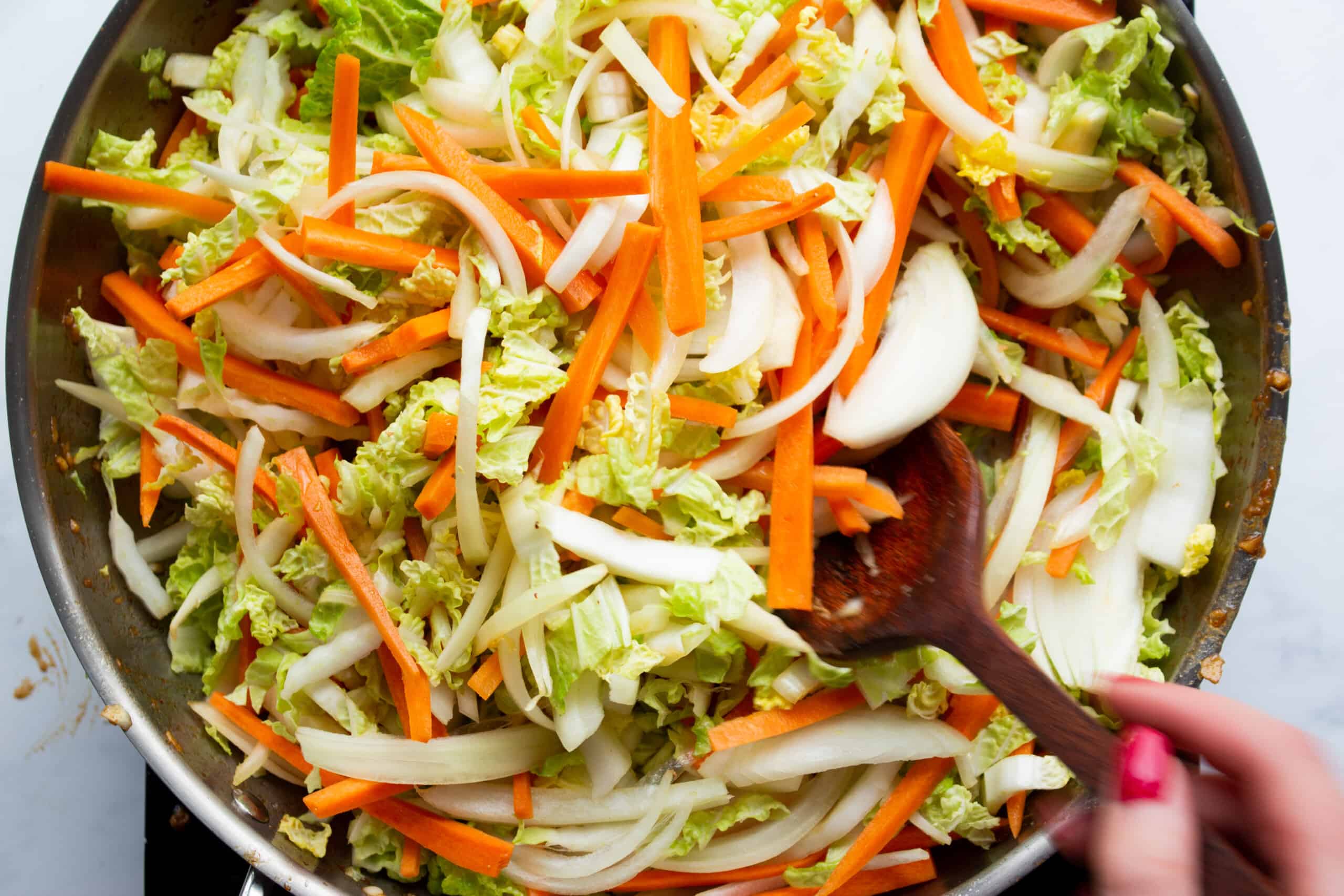 Sliced carrots and cabbage leaves in a large stainless steel pan mixed with a wooden spoon.