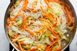 Vegetable and noodles cooked together in a large stainless steel pan on a stove.