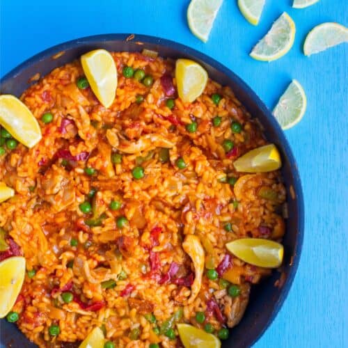 Spanish paella in a pan with decorative lemon slices scattered over and on bright blue background.