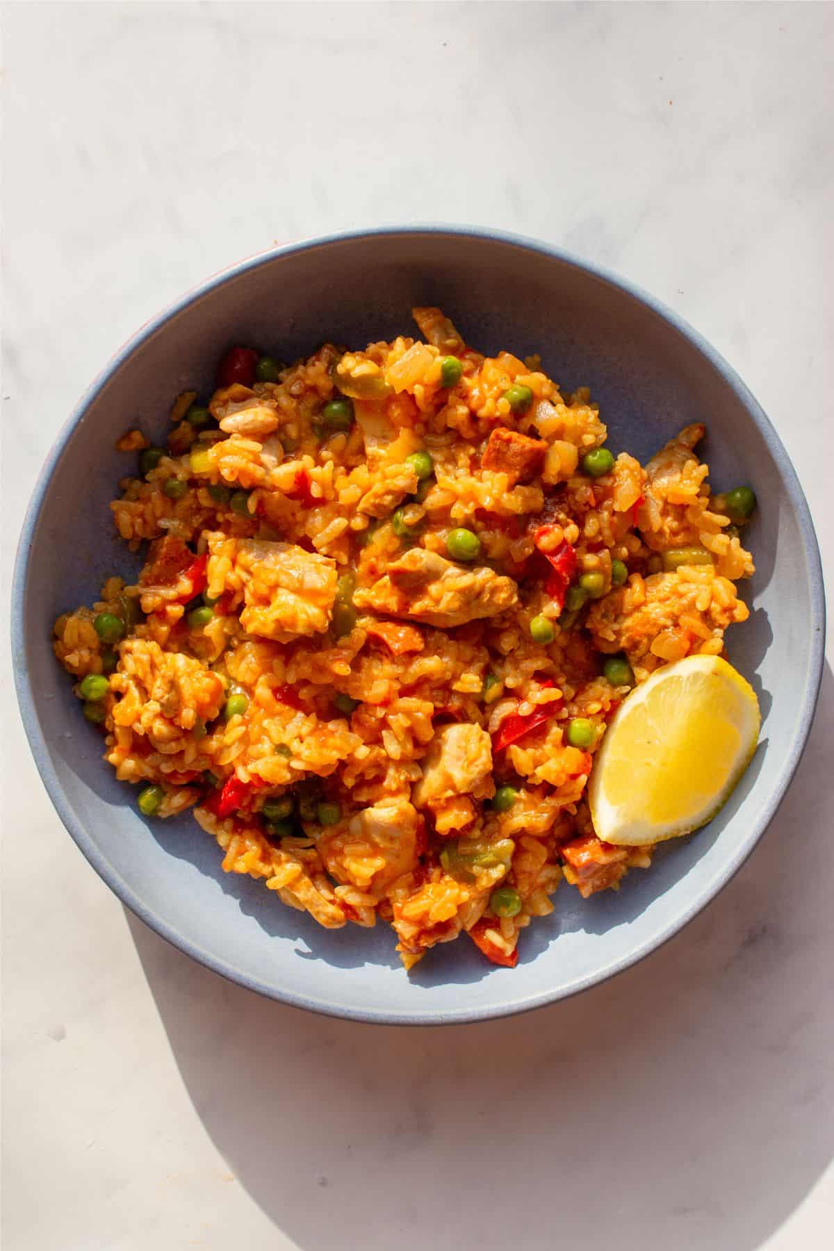 Paella rice with chorizo, chicken, peppers and peas in a bowl with a wedge of lemon.