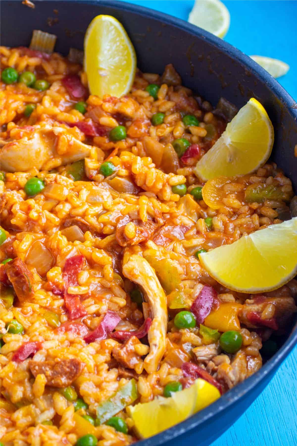 Close up of chicken paella made with rice, peppers, onions and peas with lemon wedges in the pan on a bright blue back ground.