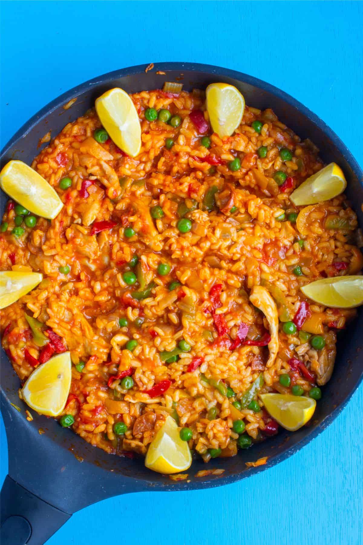 Paella served in a pan with lemon wedges decorating the edges on a bright blue background.