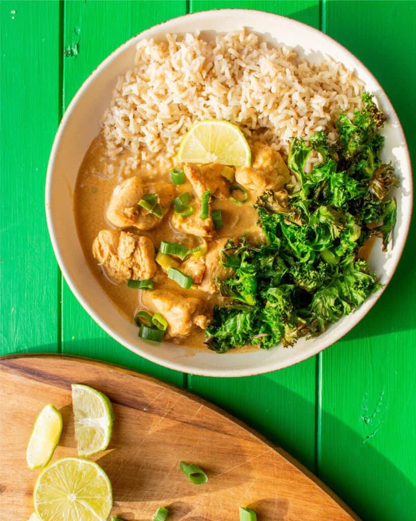 A bowl of Peanut Butter Chicken Satay Curry with brown rice, a wedge of lime and kale next to a wooden chopping board with lime wedges.