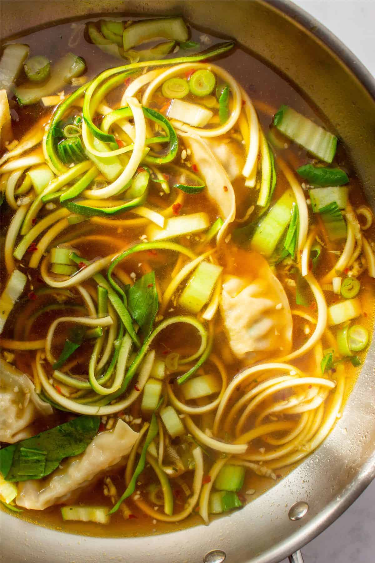 Close up of soup with dumplings, spring onions, pok choi and spiralised courgette with stock in a saucepan.