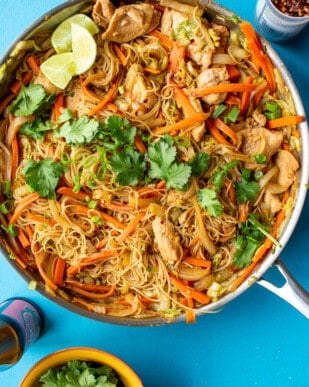 Chicken Vermicelli noodles in a large pan topped with coriander and lime wedges on a blue background.