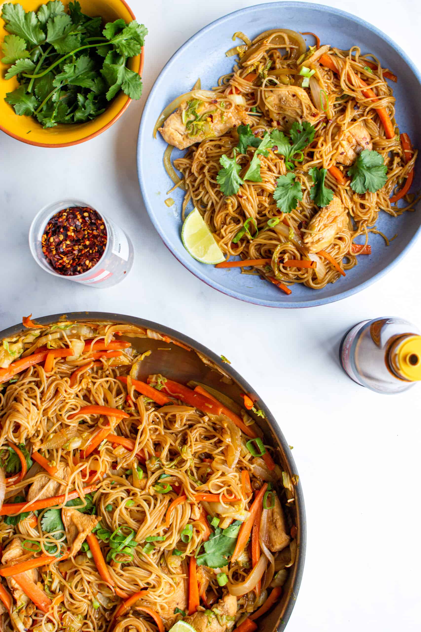 Chicken Vermicelli in a blue bow next to a pan with noodles, chicken, carrots, a container of chilli flakes and a bowl of coriander.