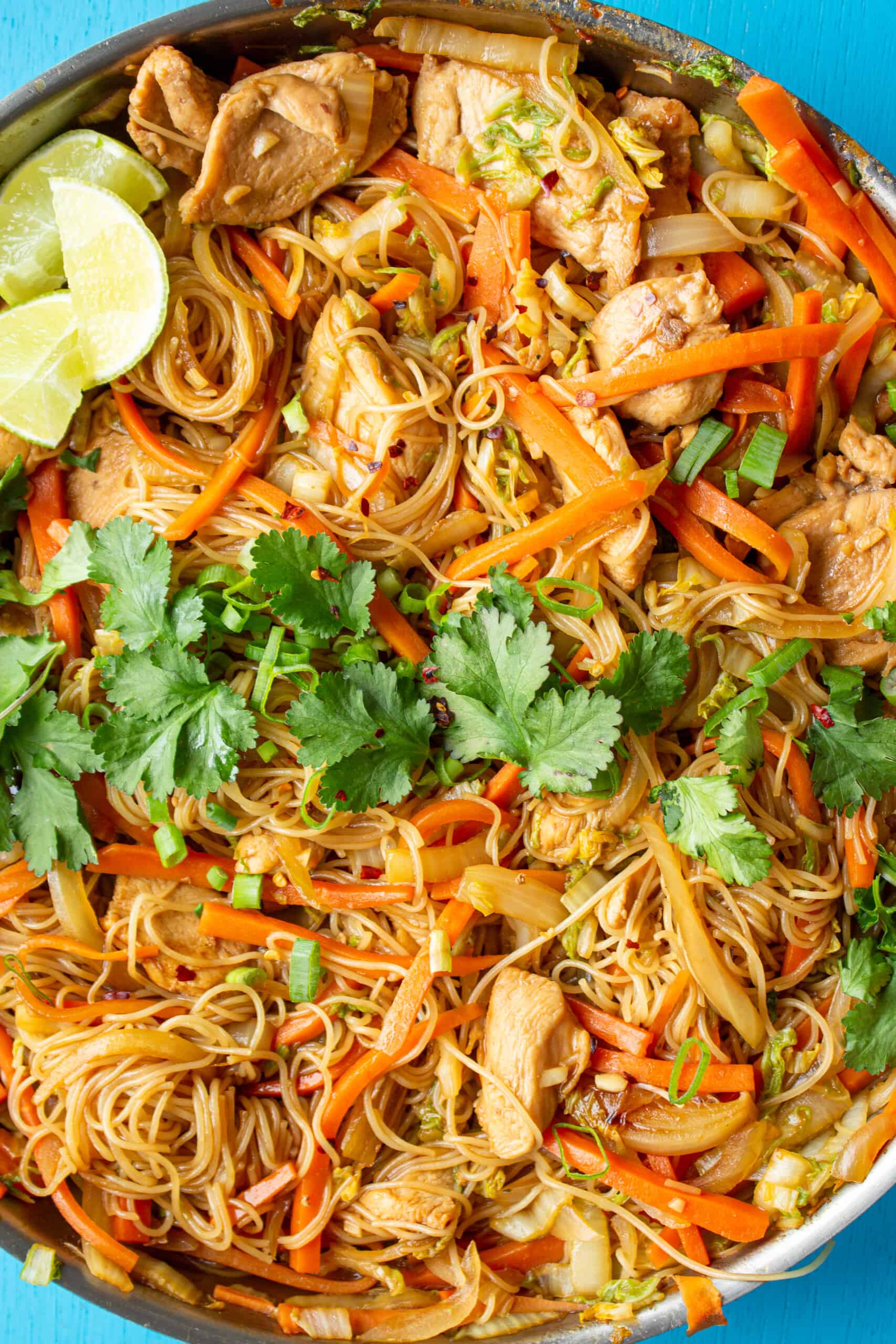 Close up of chicken vermicelli with noodles, chicken, carrots, onions, lime wedges and fresh coriander on a blue background.