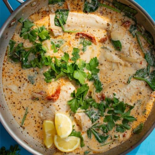 A large pan with sundried tomatoes in a creamy sauce with basa fillets topped with parsley and lemon wedges.