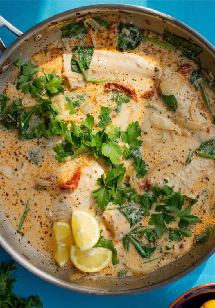 A large pan with sundried tomatoes in a creamy sauce with basa fillets topped with parsley and lemon wedges.