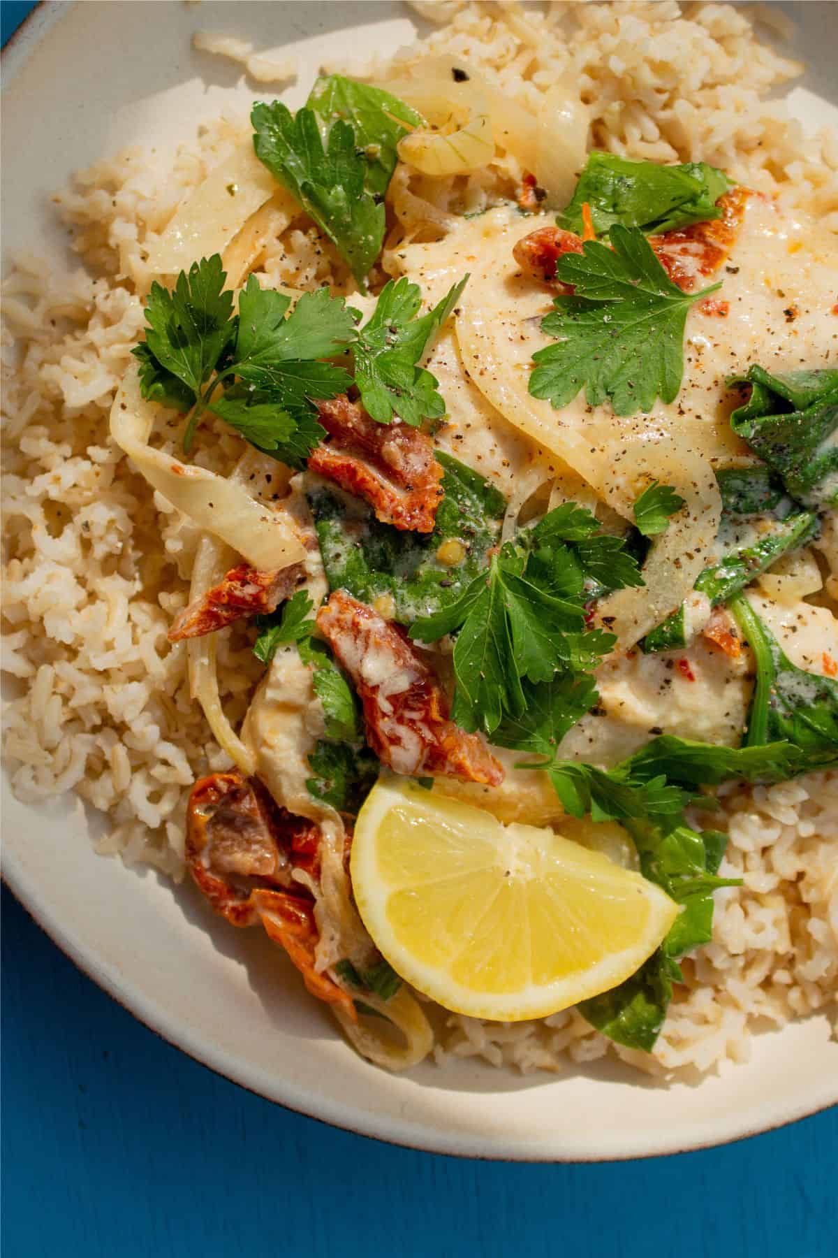 Close up of basa fillets with a creamy sauce with sundried tomatoes, served with rice, fresh parsley and a wedge of lemon.