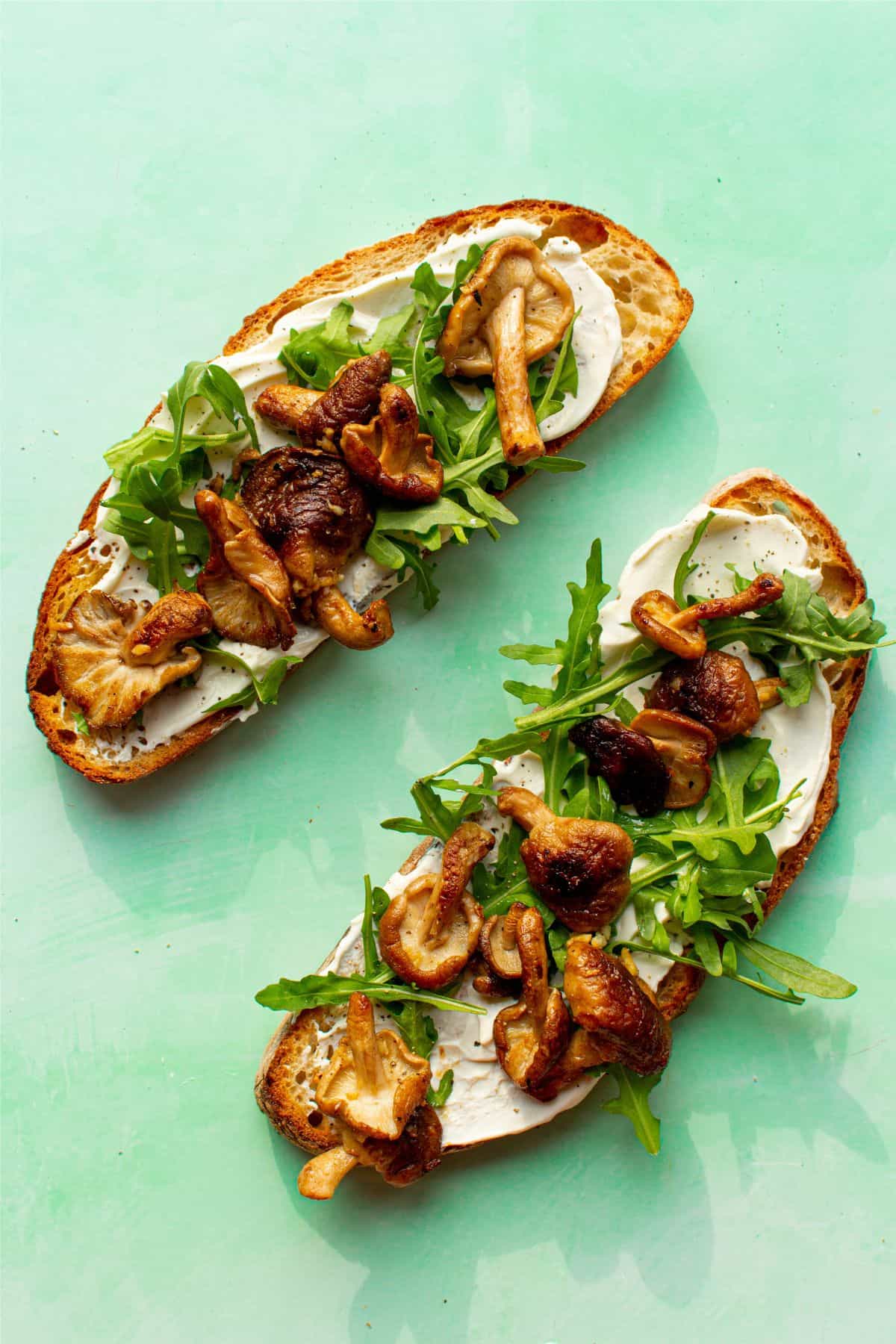 Mushroom toast with 2 pieces of sourdough, mushrooms and cream cheese and rocket on a pale green background.