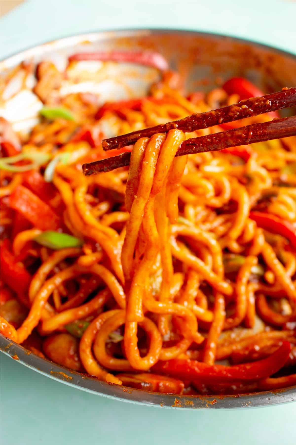 Noodles with chopsticks in pan with peppers and tomatoey red sauce in a stainless steel pan.