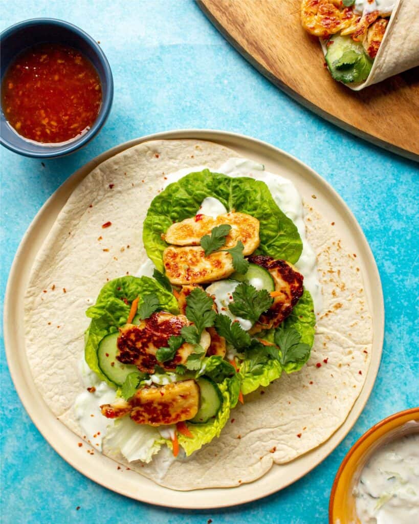 A wrap with lettuce, halloumi, tzatziki and a pot of sweet chilli sauce and another wrap on a wooden board.