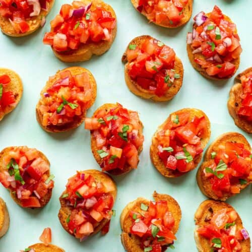 Mini bread rounds with chopped tomatoes, toasted baguette slices, red onion and basil on a pale green background.
