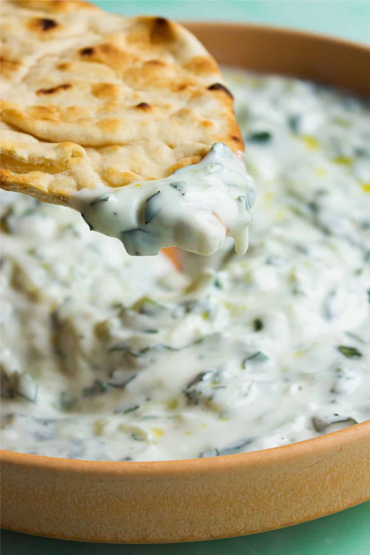 Close up of Tzatziki with a piece of flat bread being dipped into the bowl.