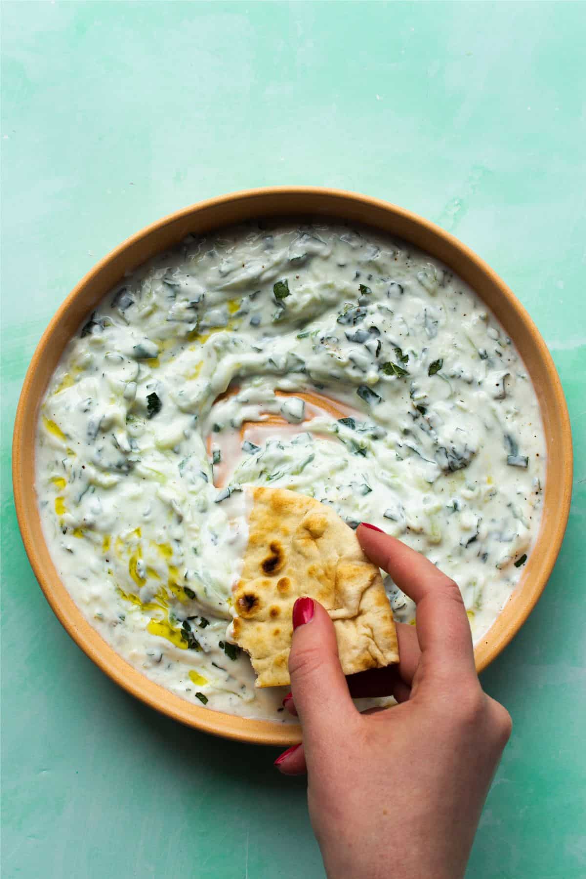 An overhead vew of a bowl of Tztziki topped with oil being dipped with part of naan bread held with hand.