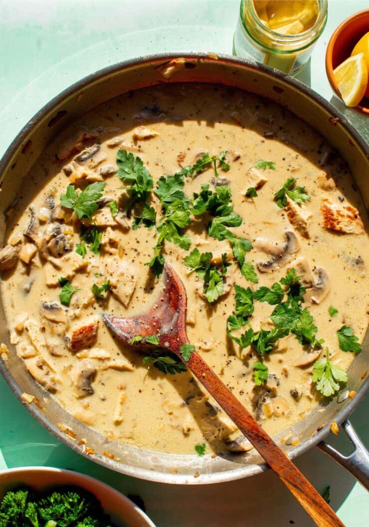 Creamy pork in sauce in a large pan with mushrooms and topped with fresh parsley and mixed with a wooden spoon.