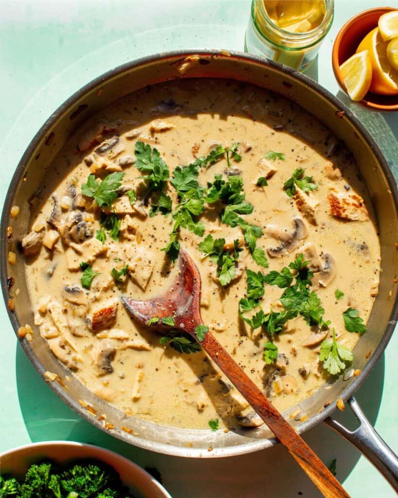Creamy pork in sauce in a large pan with mushrooms and topped with fresh parsley and mixed with a wooden spoon.