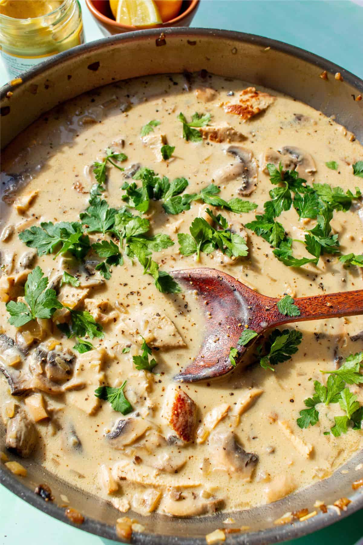 Large pan withcooked pork stroganoff with mushrooms and topped with fresh parsley and mied with a wooden spoon.