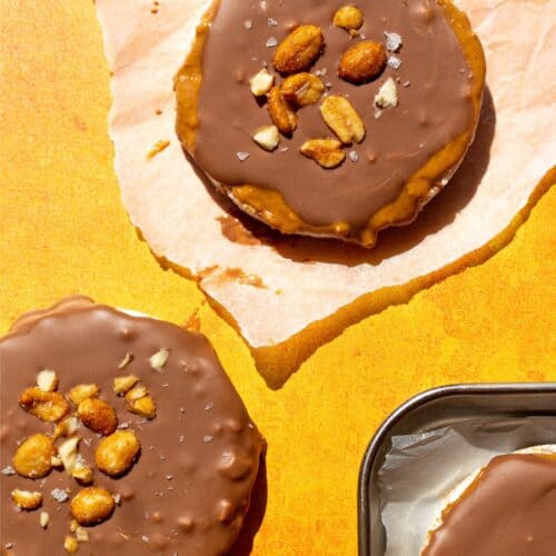 Rice cakes topped with chocolate and peanuts, one on parchment paper, the other on an orange background and a tray with more rice cakes.
