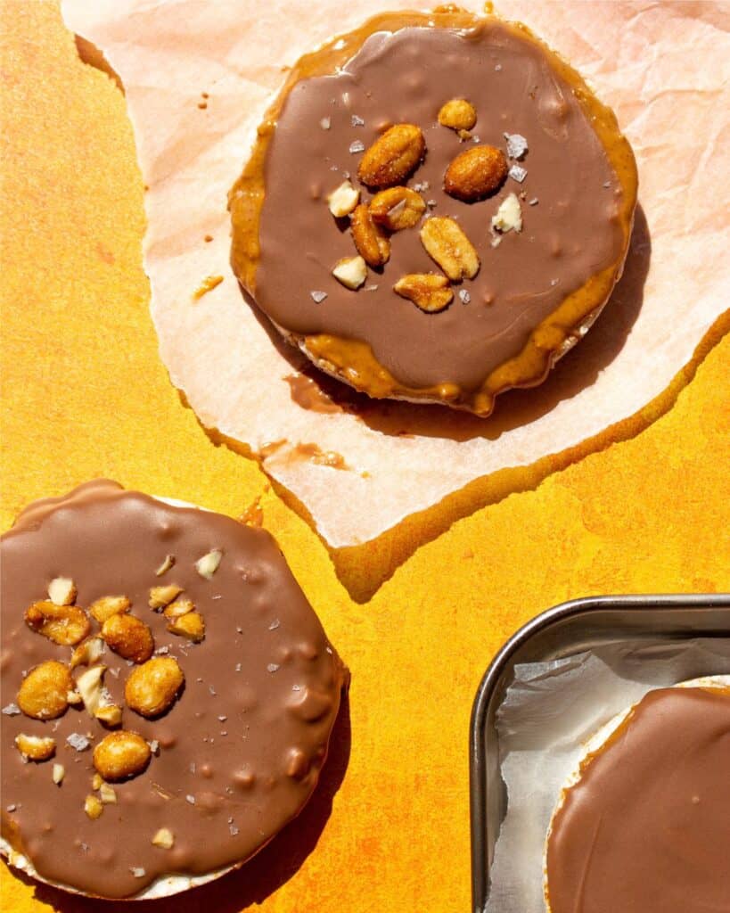 Rice cakes topped with chocolate and peanuts, one on parchment paper, the other on an orange background and a tray with more rice cakes.