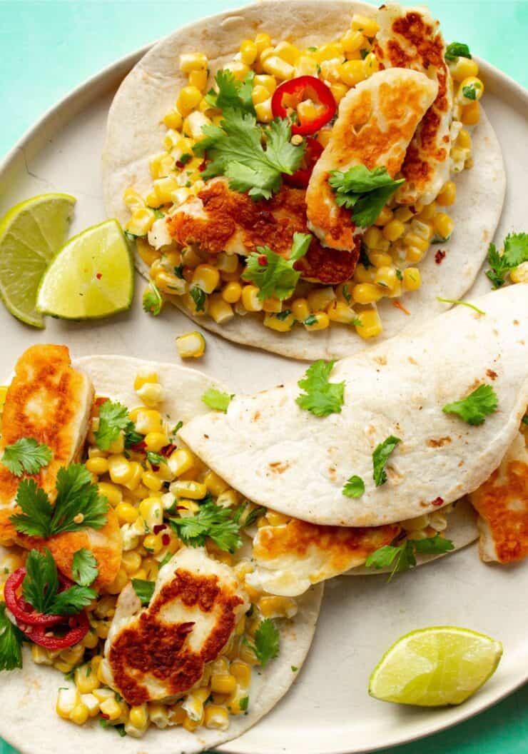 Tacos with sweet corn, golden browned halloumi, red chillies and coriander on plate with lime wedges.