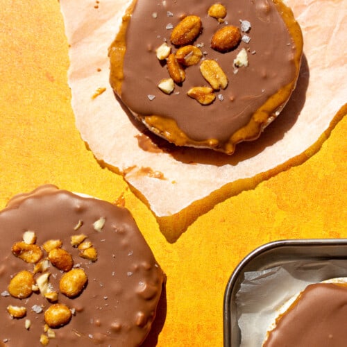 Peanut butter rice cakes (one on parchment paper and one on orange background) with chocolate topping, peanuts and flakey salt.