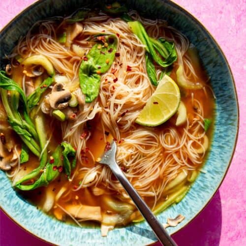 Noodles in a blue bowl with a pink background with noodles, miso stock, pok choi, mushrooms, spring onion slices and a wedge of lime.