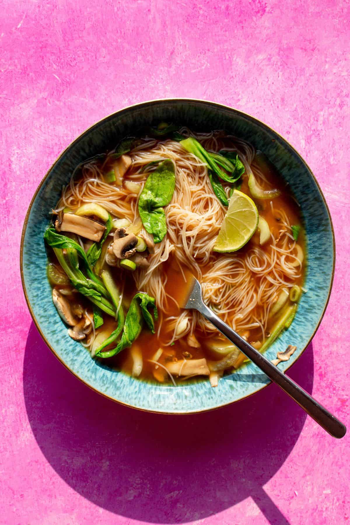 Healthy Instant noodles in a blue bowl with a pink background with noodles, miso stock, pok choi, mushrooms, spring onion slices and a wedge of lime.
