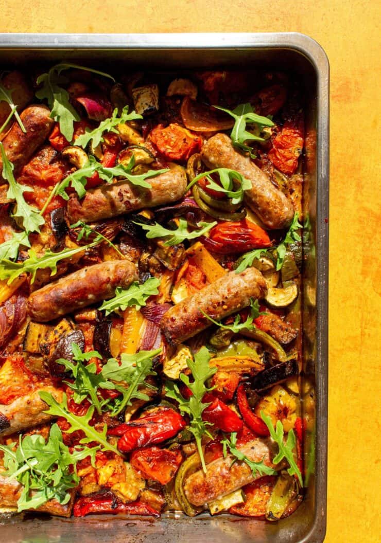 Roasted vegetables and sausages in a tray garnished with rocket on a yellow background.