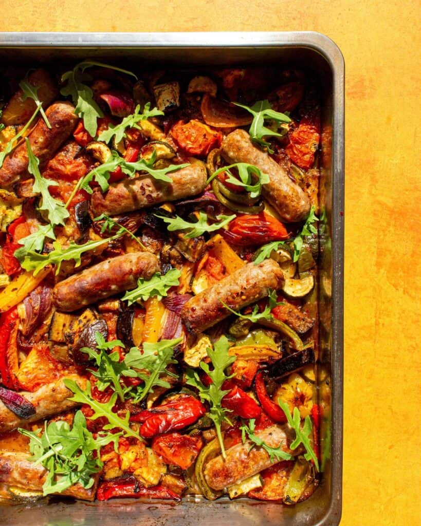 Roasted vegetables and sausages in a tray garnished with rocket on a yellow background.
