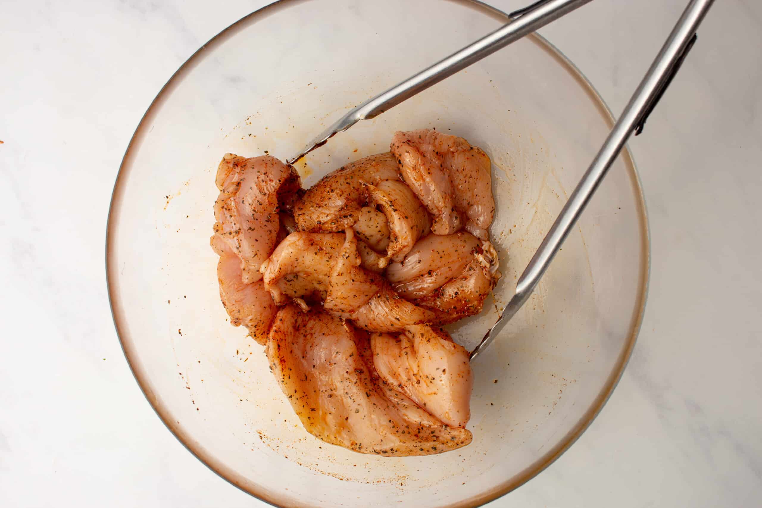 Chicken breast pieces in a glass bowl coated with Cajun spices and mixed with metal tongs.