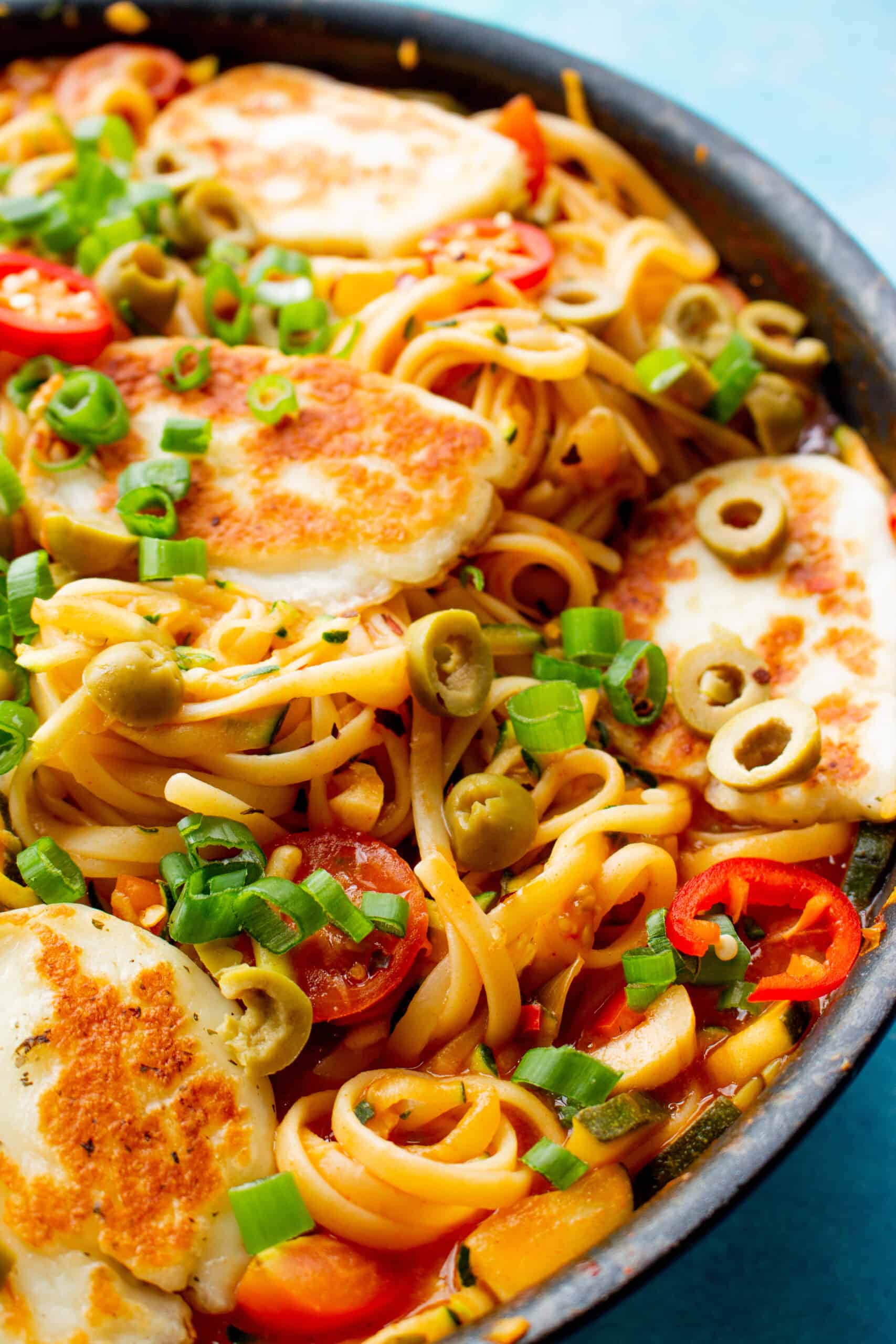 Close up of spaghetti, slices of spring onion, red chillies and golden browned halloumi in a pan.