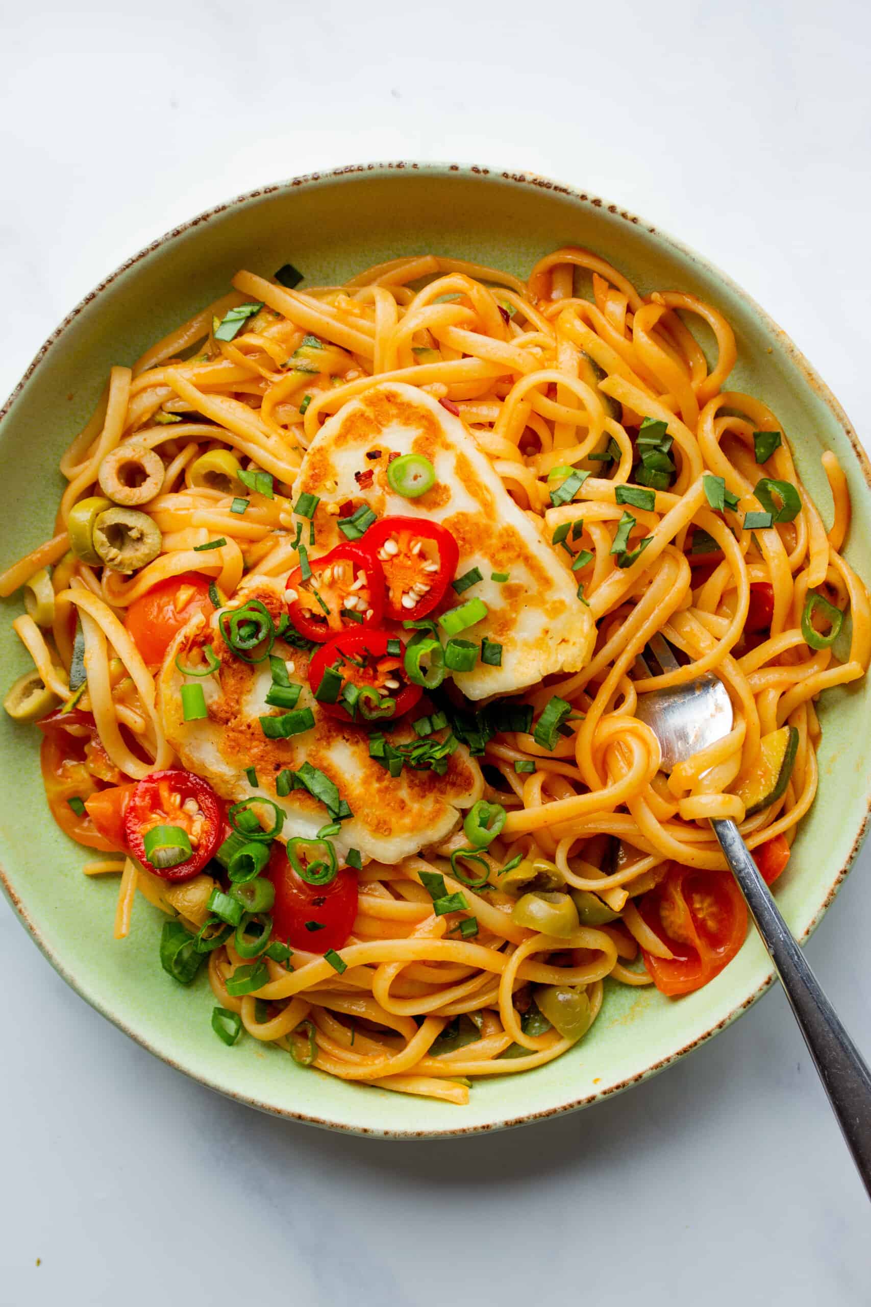 Halloumi pasta served in a pale green bowl with golden browned halloumi, slices of spring onion, chillies and tomatoes with a fork.
