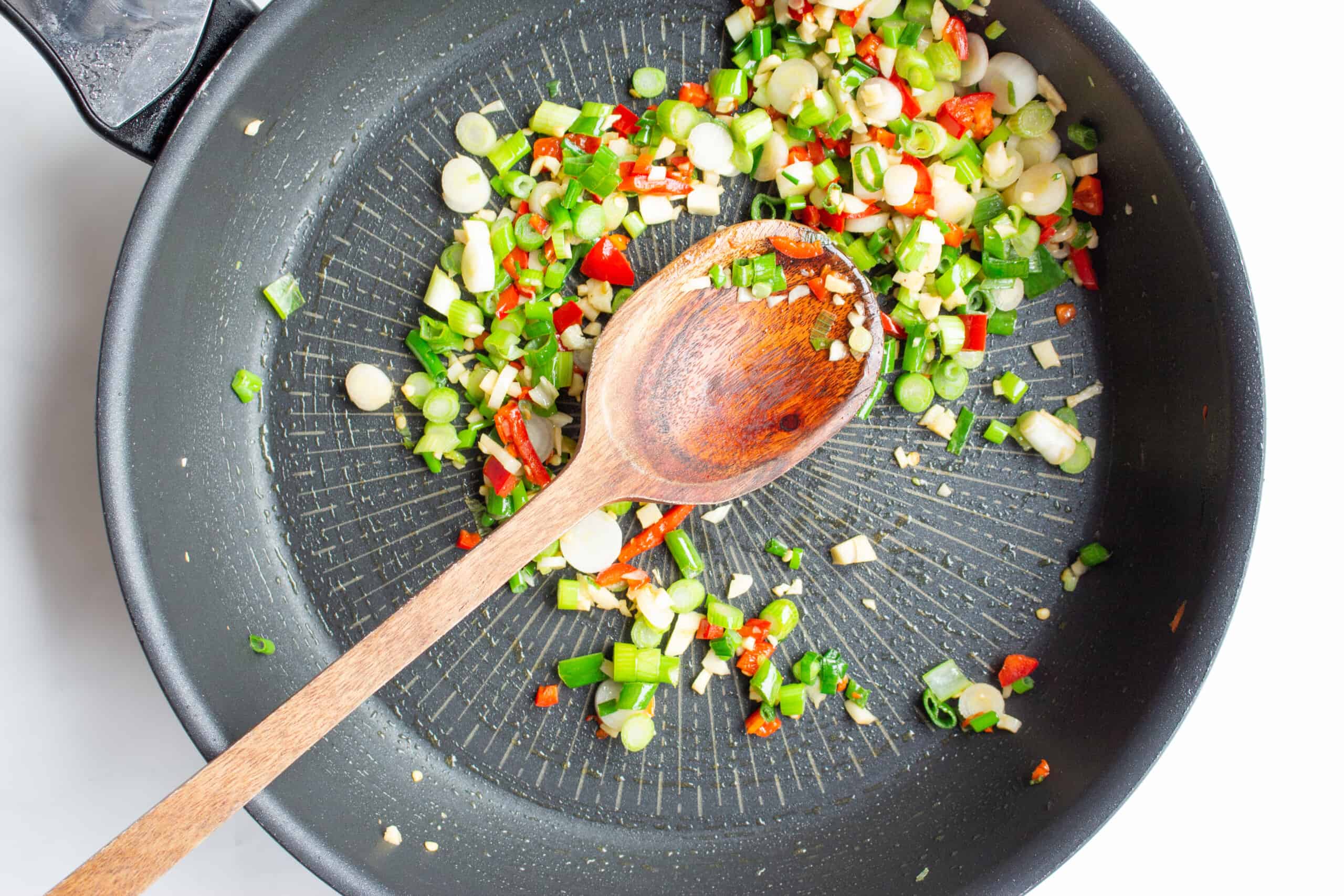 Slices of chillies, garlic and spring onions in a black pan with a wooden spoon.