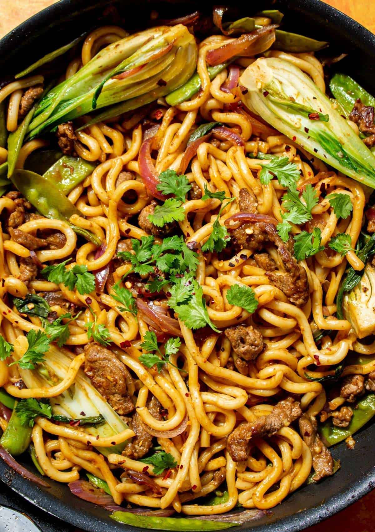 close up shot of udon noodles with beef strips in the pan with pak choi, red onion and coriander.