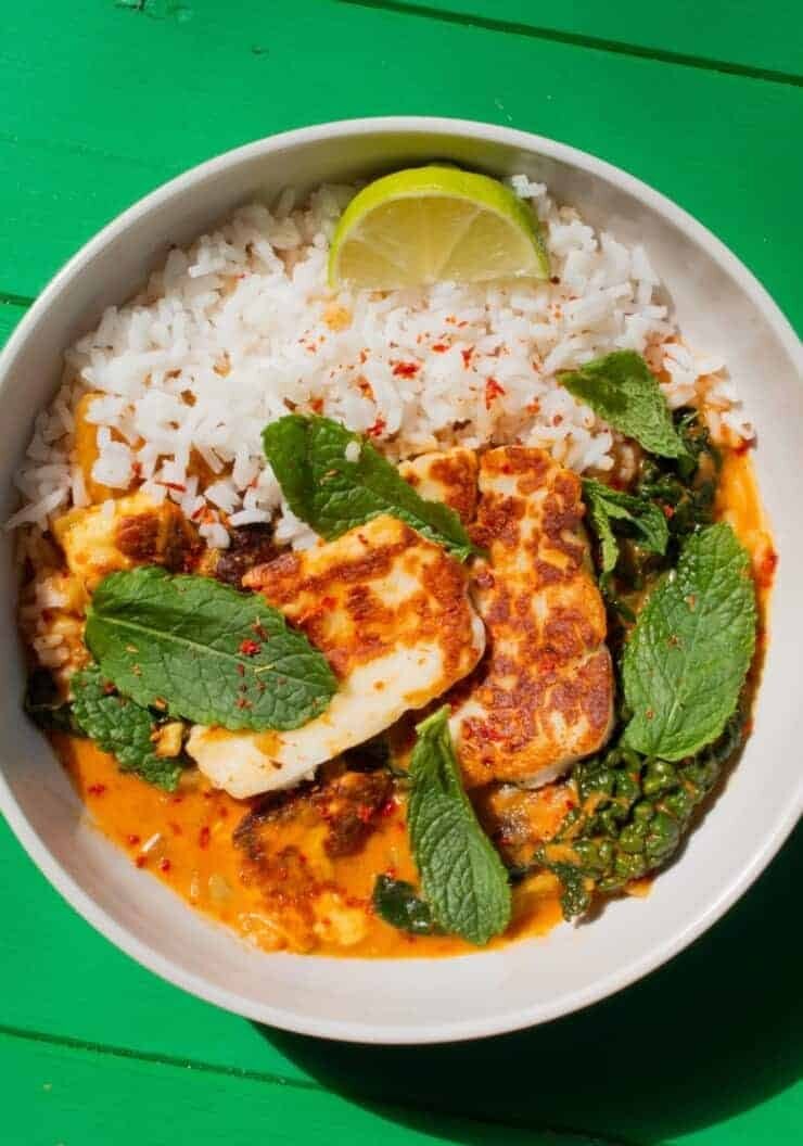 Halloumi curry in a white bowl with rice, fresh mint and a wedge of lime on a green background.