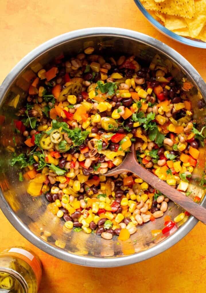 Cowboy caviar in a large metal bowl with sweet corn, mixed beans, coriander and jalapenos with a wooden spoon on an orange background.