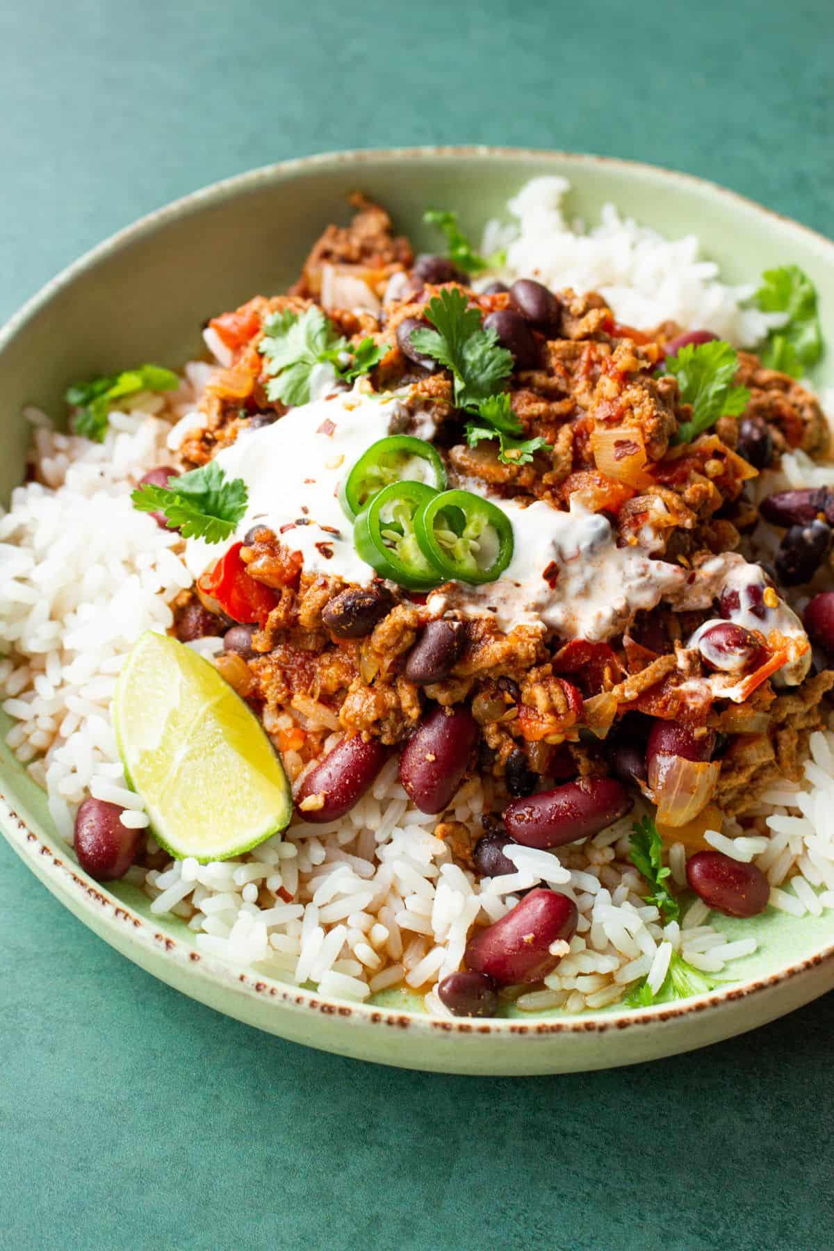 Close up of a bowl of slow cooker chilli con carne, garnished with wedge of lime, green chilli slices, coriander and sour cream.