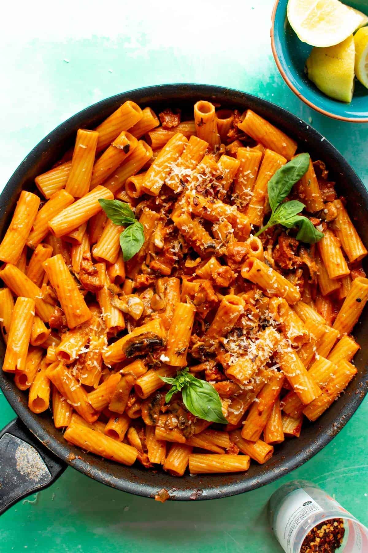 Creamy Chorizo pasta with basil and parmesan in large pan on a green back ground, with lemon wedges in bowl and chilli flakes in container. 