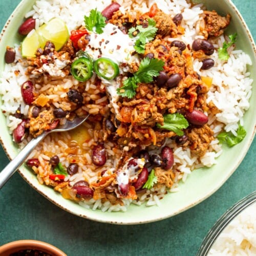 Slow cooker chilli con carne on a bowl on a bed of rice garnished with coriander and green chillies, partial view of chilli flakes and rice.