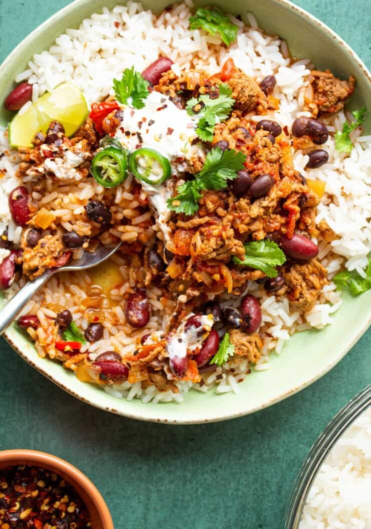 Slow cooker chilli con carne on a bowl on a bed of rice garnished with coriander and green chillies, partial view of chilli flakes and rice.