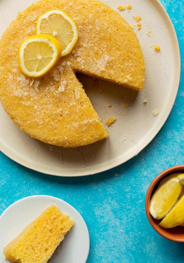Steamed Lemon Cake (with slices on lemon on top) on a large plate with a slice cut out and served on a plate next to a bowl of lemon wedges in bowl.