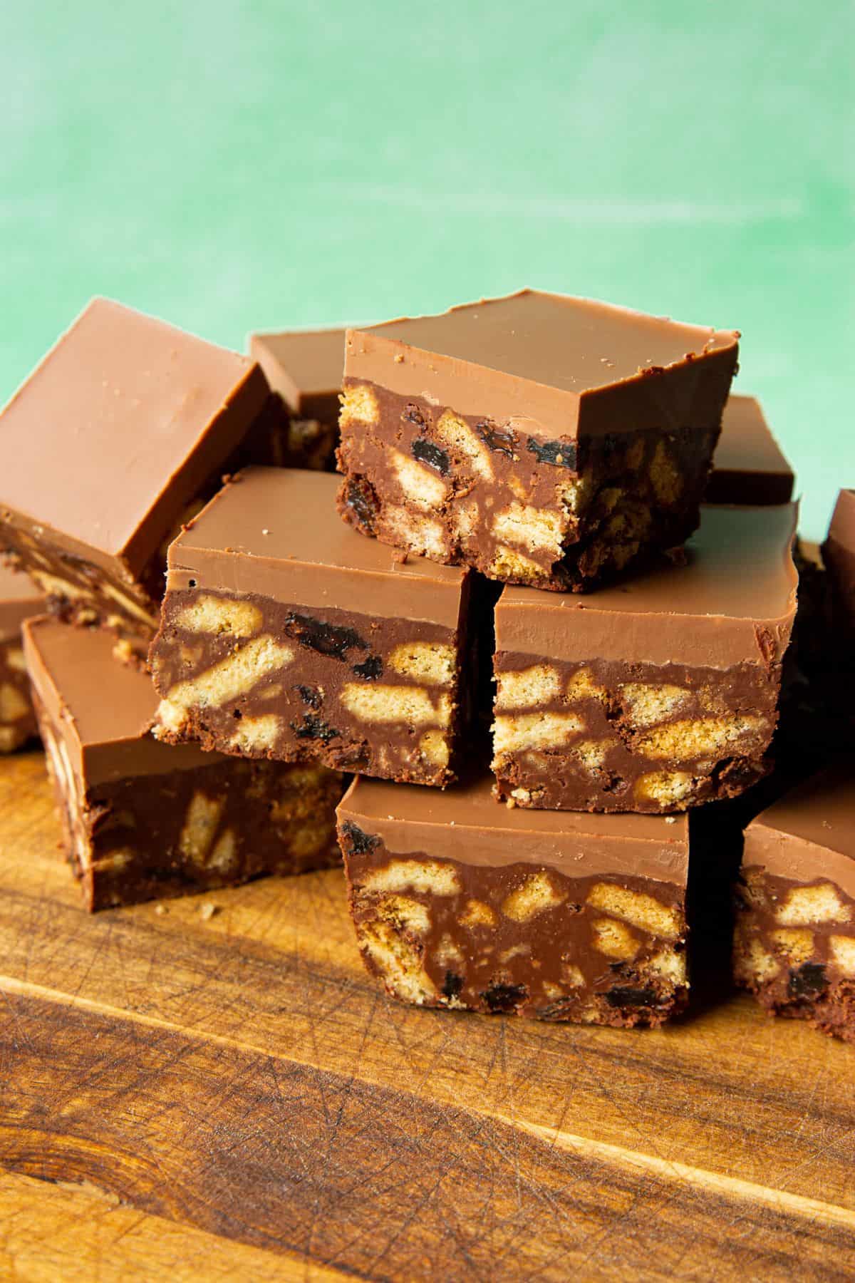 Tiffin Cake square slices, containing biscuit, raisins and thick chocolate topping stacked on top of each other on a chopping board.